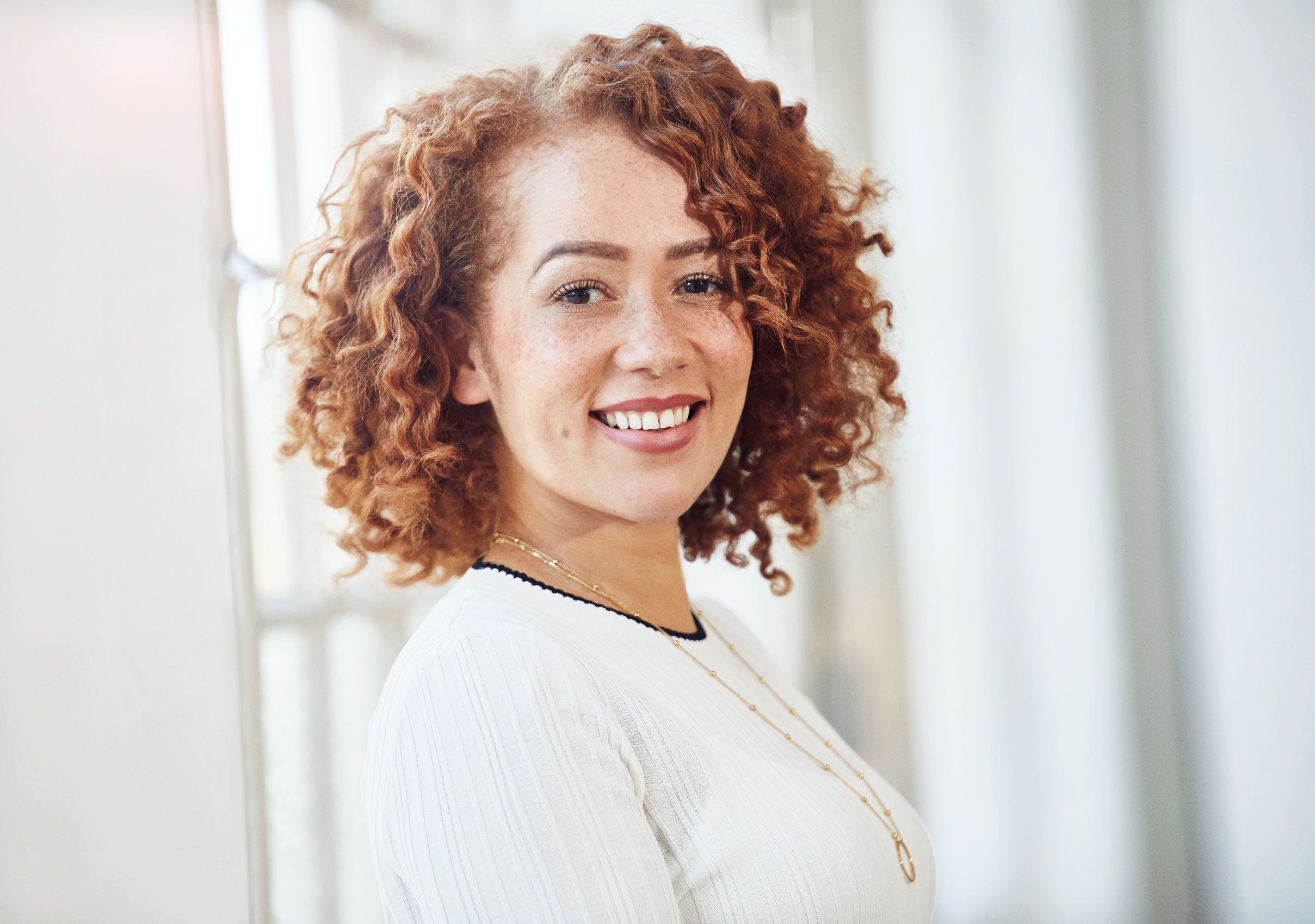 Corporate Portraits - Woman with curly hair standing by office windows smiling - shot for adverting and marketing based company.  London