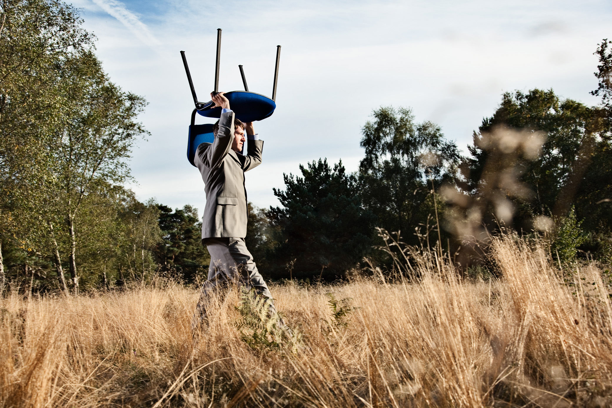 Concept imagery - business man walking through field carrying office chair.  Shot on location in Sussex, UK
